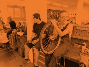 Auszubildende sitzen auf einer Werkbank in einer Fahrradwerkstatt. Einer hält ein Laufrad. Das Bild ist orange eingefärbt.