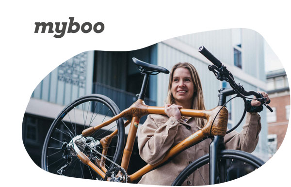 MyBoo Markenvorschau - Eine Frau trägt ihr Bambus-Fahrrad. Sie hat einen beigen Mantel. Darüber ist das Myboo-Logo abgedruckt.