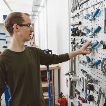 Ein junger Mitarbeitender vom Team von FahrRad R18 sucht Werkzeug (blau) von einer Werkzeugwand heraus. Er trägt einen schwarzen Pullover und eine Brille.