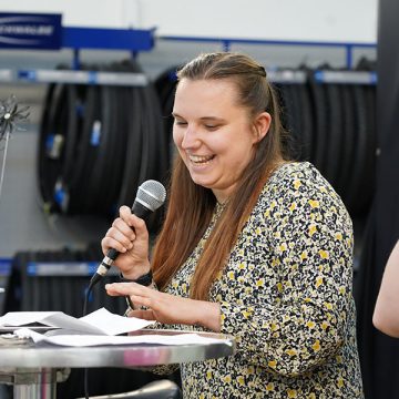 Eine Rednerin im Kleid mit Blümchenmuster und mit braunen Haaren spricht an einem Stehtisch in ein Mikrofon