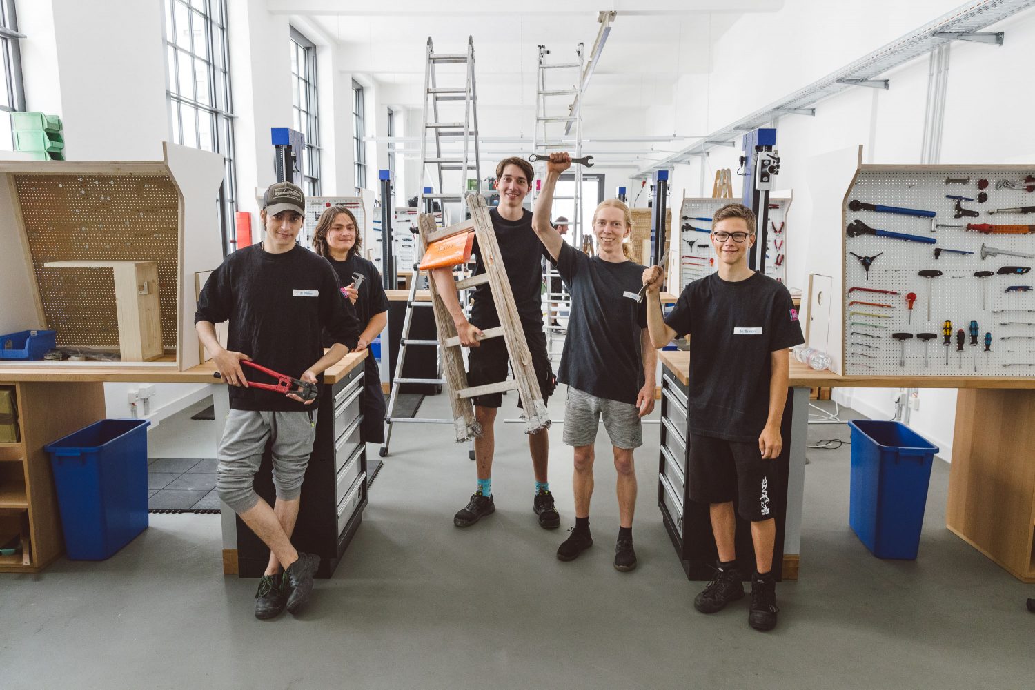Ein Team aus jungen, männlichen Mitarbeitern in der Werkstatt von FahrRad R18. Sie tragen schwarze Tshirts mit Firmierung und wirken Motiviert. Einige halten Werkzeug (in die Luft) und einer steht neben einer Tretleiter.