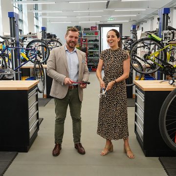 Ein Mann und eine Frau stehen mit Klemmzangen in der Werkstatt von FahrRad R18. Anlässlich der Eröffnung haben Sie mit den Geräten ein Fahrradketten-Absperrband durchtrennt (nicht im Bild). Sie tragen Anzug und Kleid.